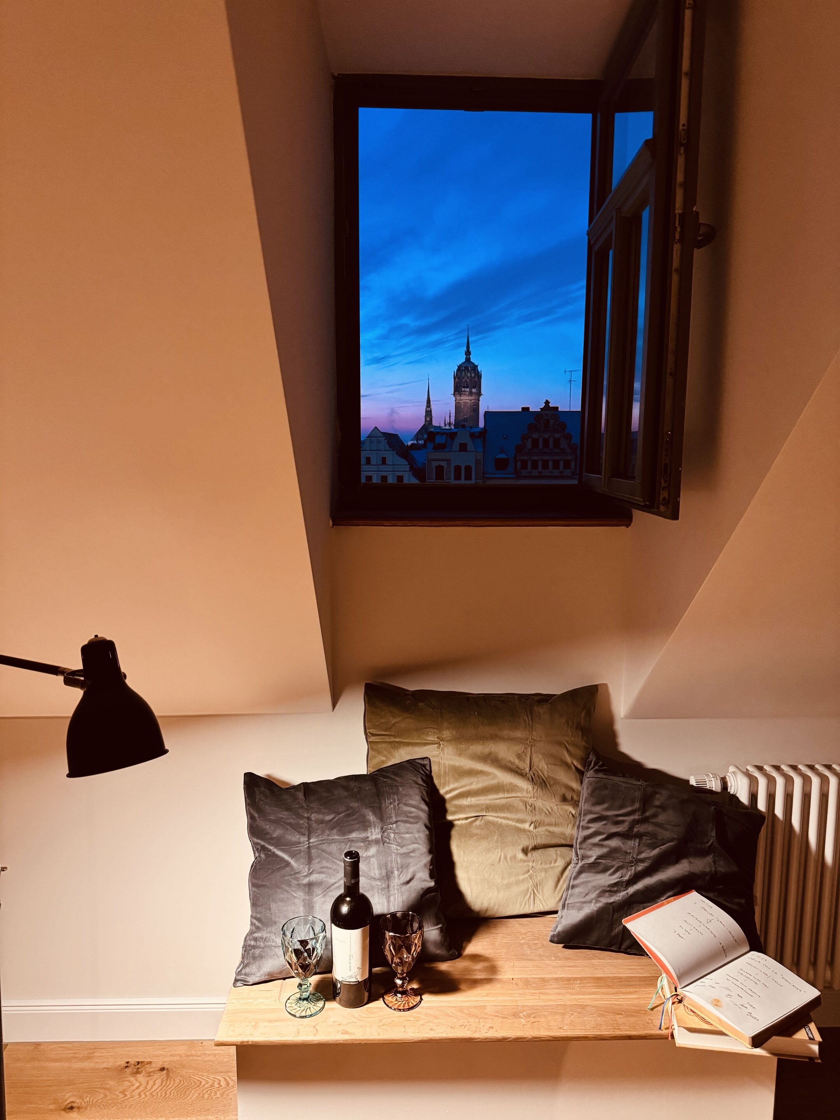 A cozy room with a sloped ceiling, a window overlooking a cityscape, a bedside table with a bottle, glasses, and an open book.