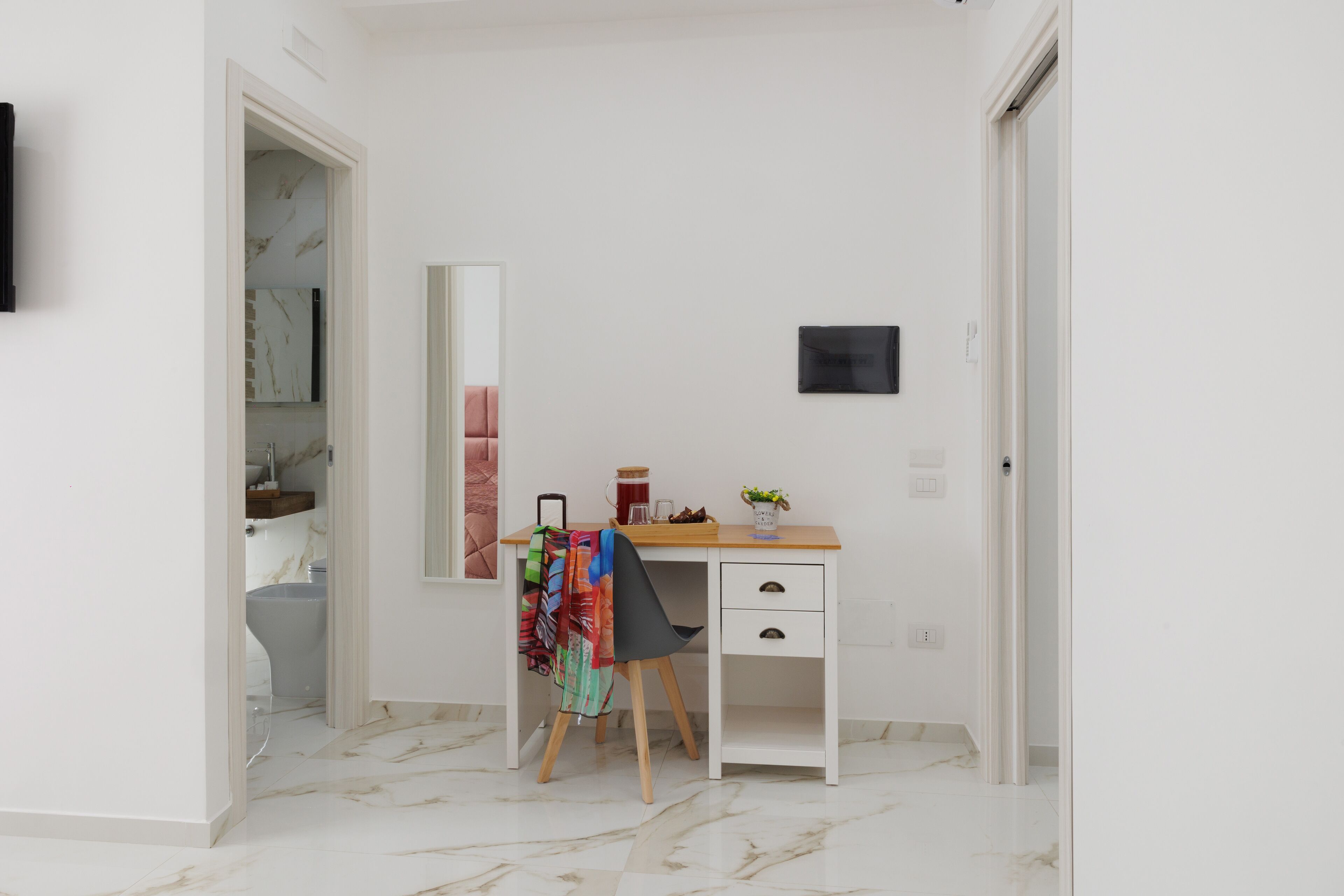 A white room with a desk, chair, and a mirror. There is a toilet visible through an open door.