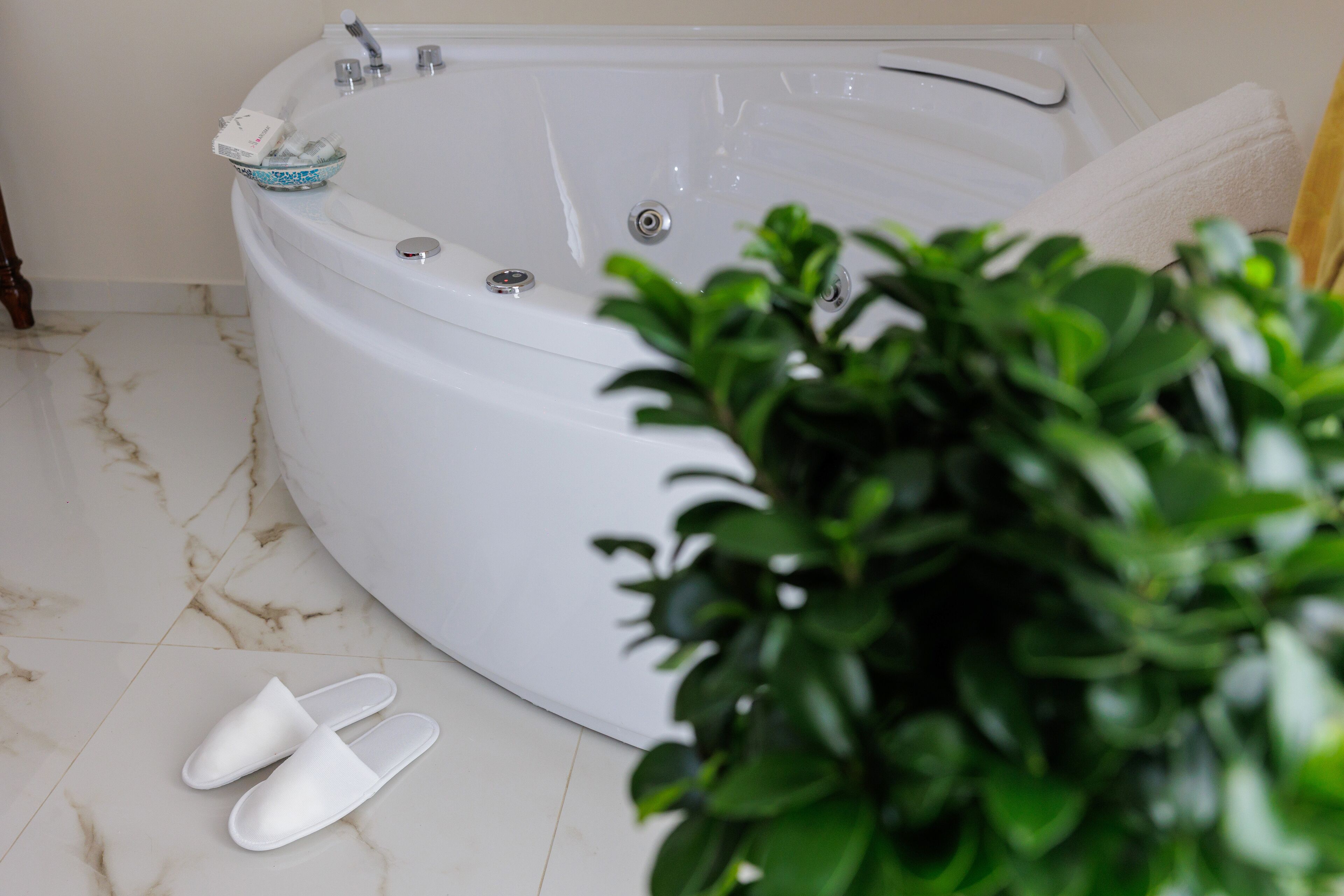 A white bathtub with a glass bowl, a white towel, and a pair of white slippers on a marble floor.