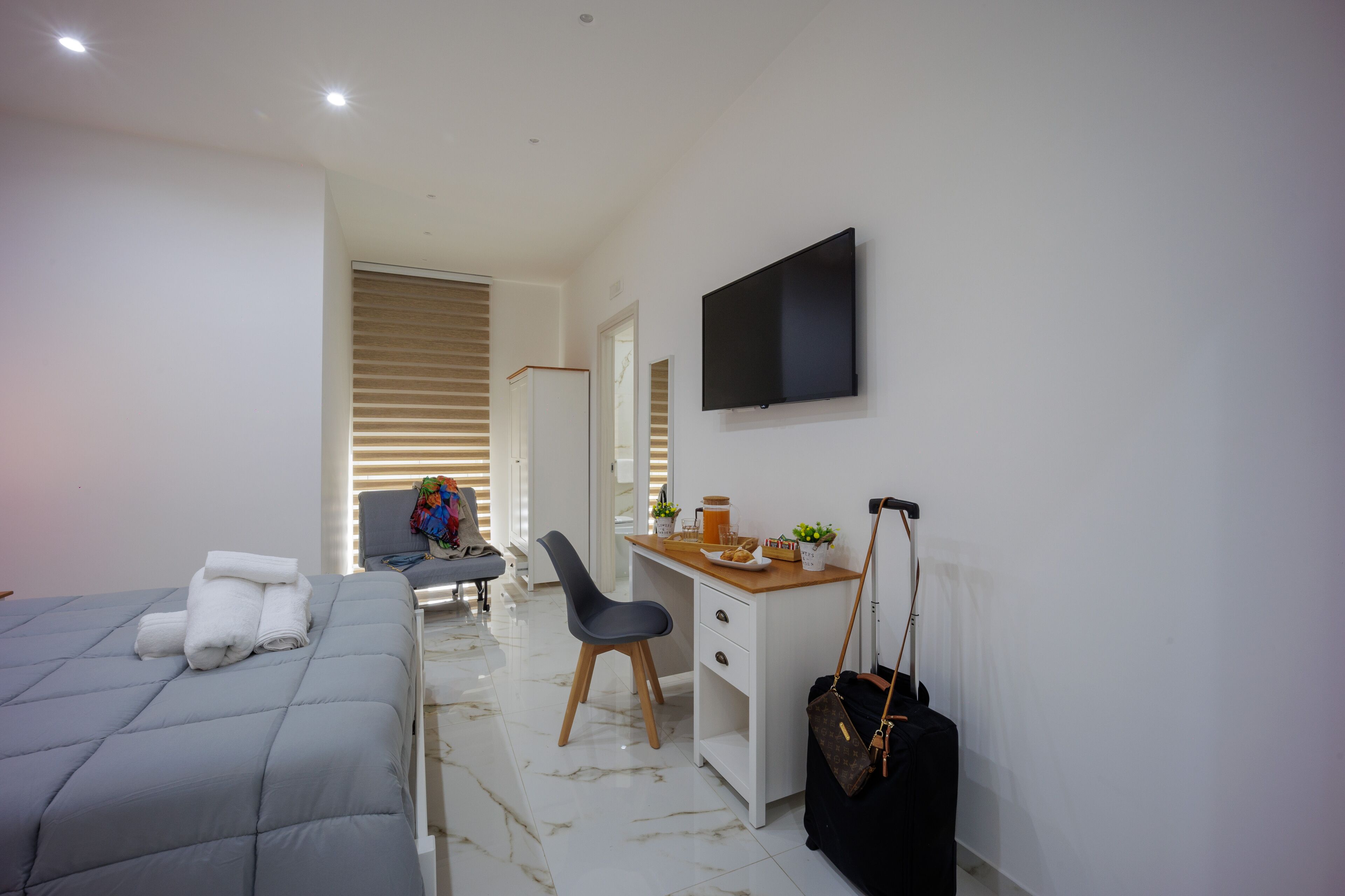 A modern hotel room with a bed, a desk, a chair, a TV, and a suitcase.
