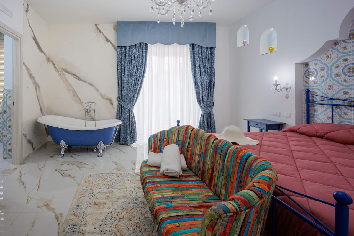 A bedroom with a large bed, a colorful sofa, a bathtub, and a chandelier.