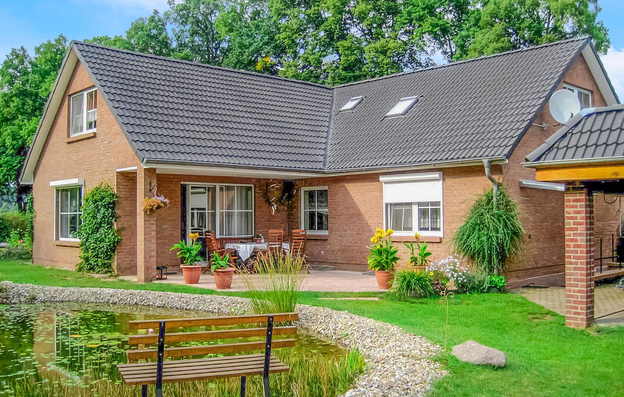 Une maison à deux étages avec un toit en pente, une véranda à l’avant avec une table et des chaises, un petit étang et un banc.