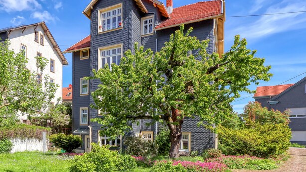 A multi-story building with a red-tiled roof, a large tree in the front yard, and a well-maintained garden with flowers. - Ferienwohnung Friedrichroda (Friedrichroda)