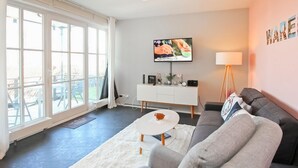 Ein Wohnzimmer mit grauem Sofa, einem weißen Couchtisch und einem an der Wand befestigten Fernseher. Es gibt ein Fenster mit Vorhängen, eine weiße Kommode mit Dekoration und eine Stehlampe.