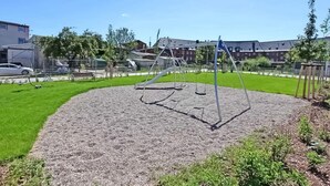 Leikkikenttä, jossa on keinut ja hiekkalaatikko, ja sen ympärillä on nurmikkoaluetta sekä taustalla näkyviä rakennuksia.
