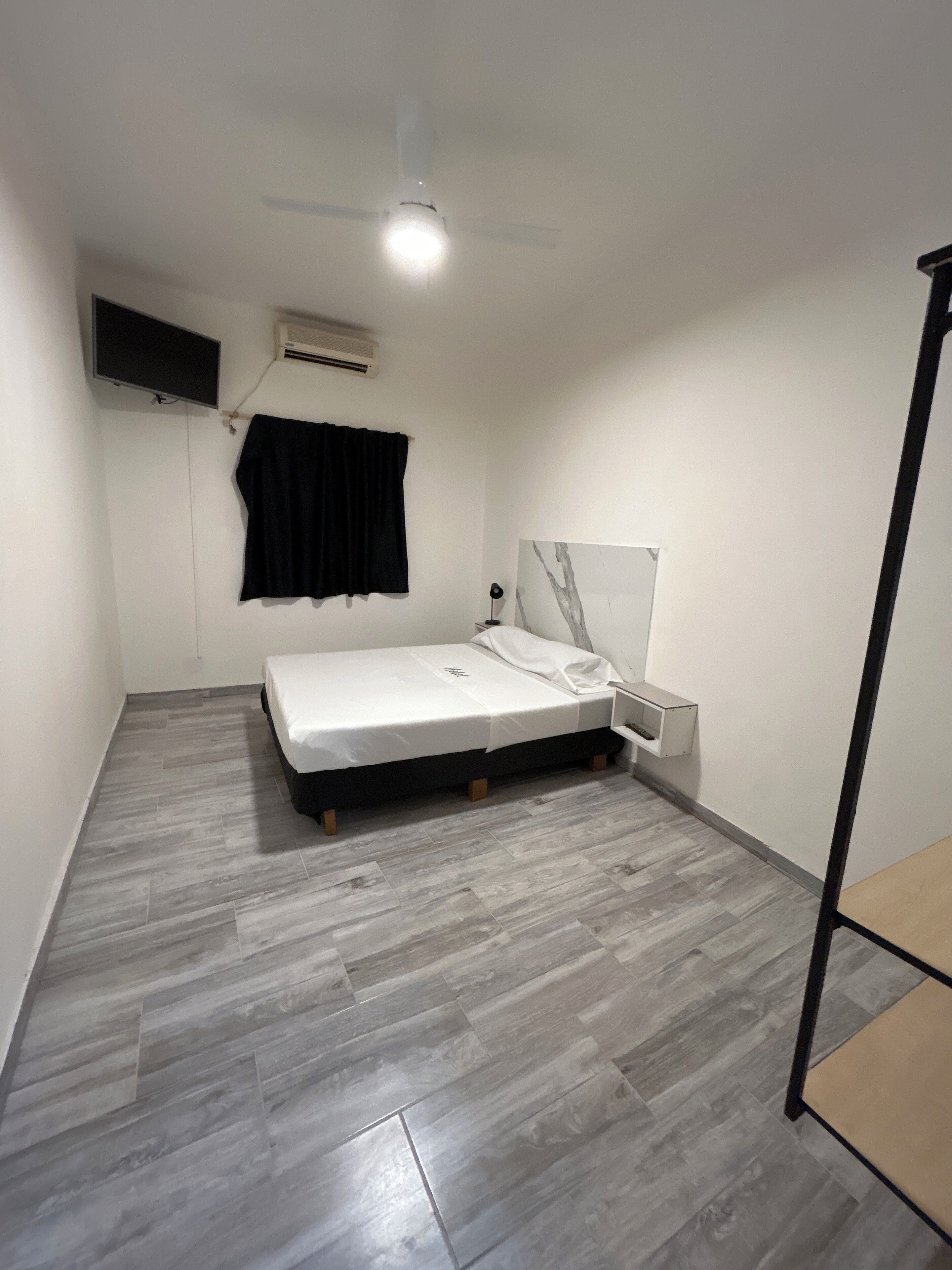 A small, modern bedroom with a bed, a nightstand, a wall-mounted TV, and a ceiling fan.