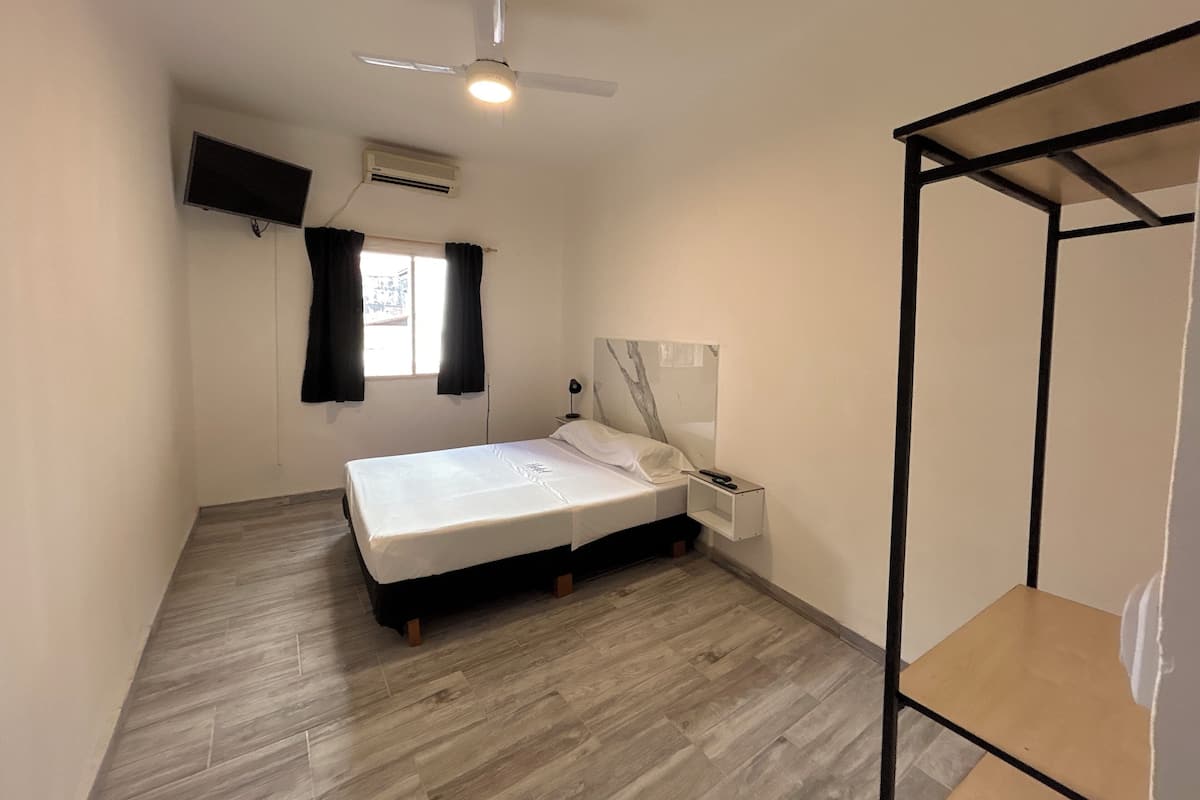 A bedroom with a bed, a nightstand, a ceiling fan, a window with curtains, and a TV mounted on the wall.