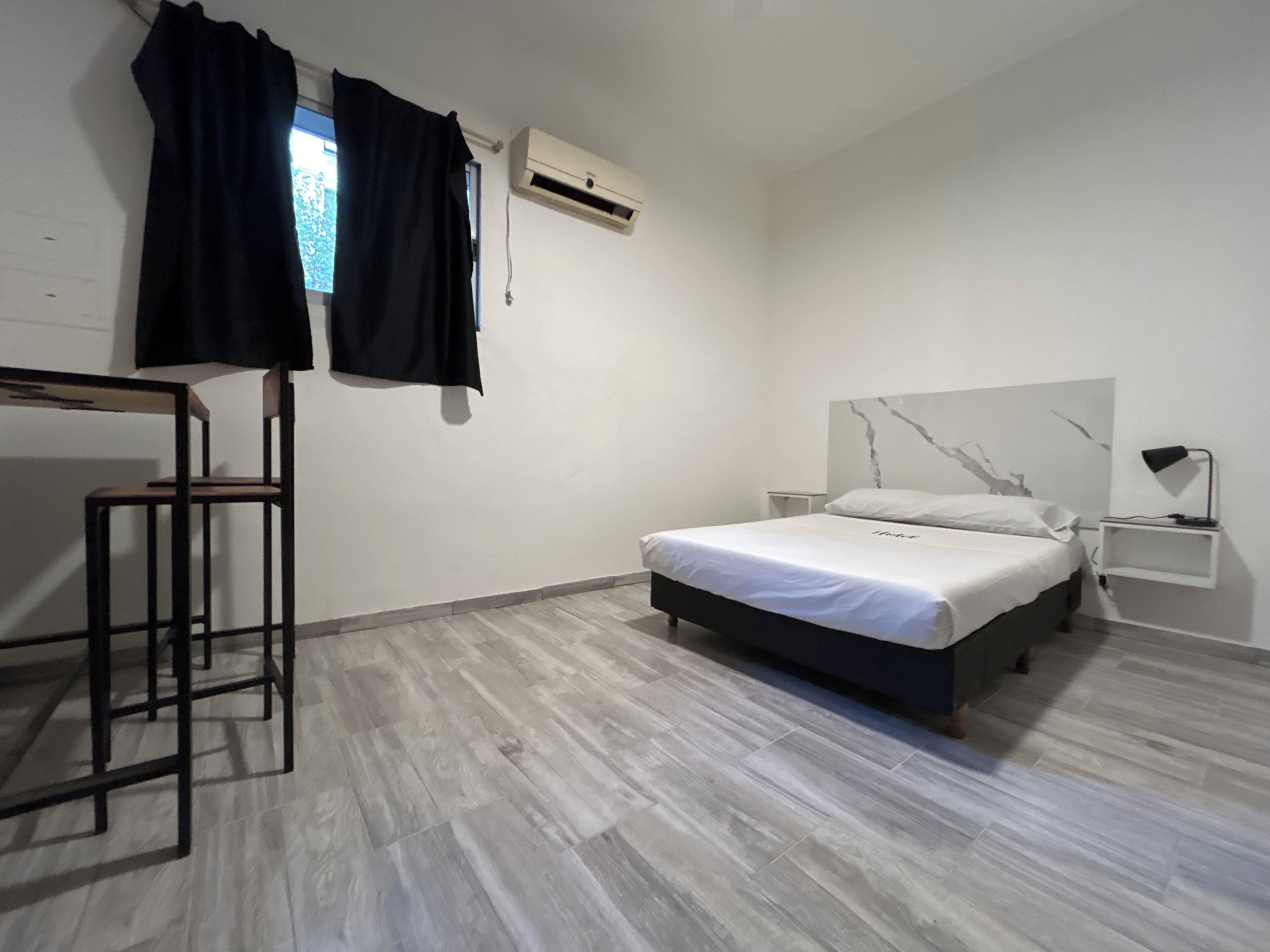 Un dormitorio con cama, escritorio y silla. Hay un equipo de aire acondicionado en la pared y una mesita de noche con lámpara.