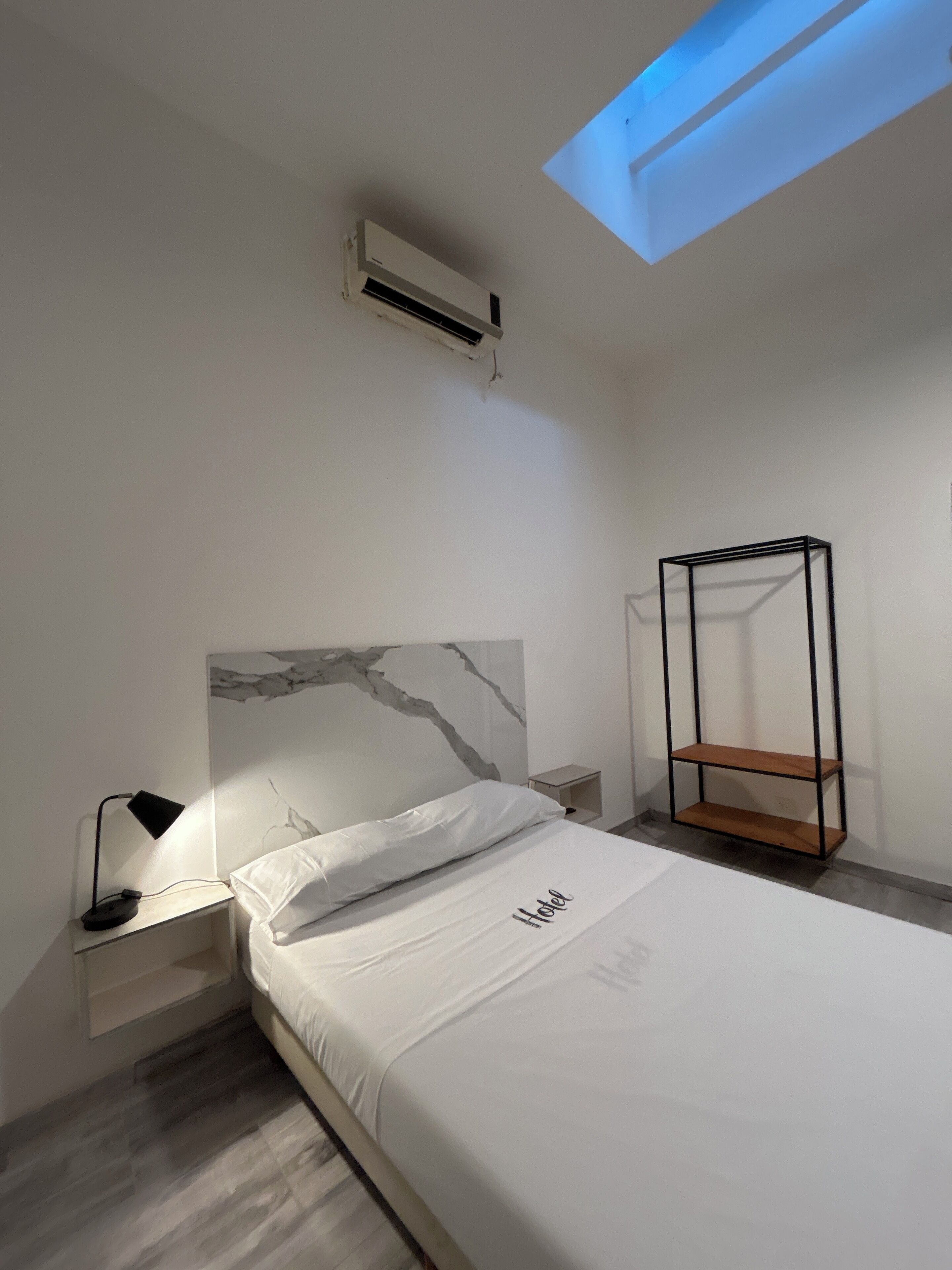 A modern hotel room with a bed, a nightstand with a lamp, a wooden shelf, and an air conditioning unit.