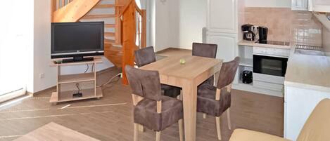 Un espacio de estar moderno con una escalera de madera, una zona de comedor con mesa y sillas, un televisor sobre un mueble bajo y una kitchenette con cafetera.