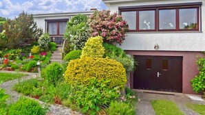 Une maison moderne à deux étages, dotée d’un jardin bien entretenu, d’un escalier apparent et d’une porte de garage.