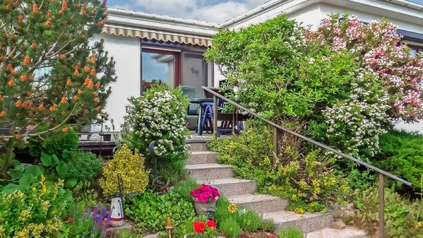 Un jardin bien entretenu, agrémenté de diverses plantes et fleurs, mène à une maison dont on aperçoit l’escalier.