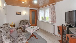 Ein Wohnzimmer mit einer Couch, einem Sessel, einem kleinen Tisch mit einer blumigen Tischdecke, einem Fernseher und Lautsprechern.
