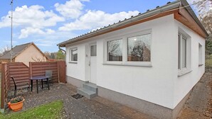 Ein kleines, eingeschossiges Haus mit weißer Fassade, einem Holzzaun, einer Terrasse mit Tisch und Stühlen und blauem Himmel.
