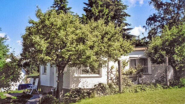 Ein Haus mit umzäuntem Garten, einem Weg zu einer Sitzgelegenheit und einem Baum davor.