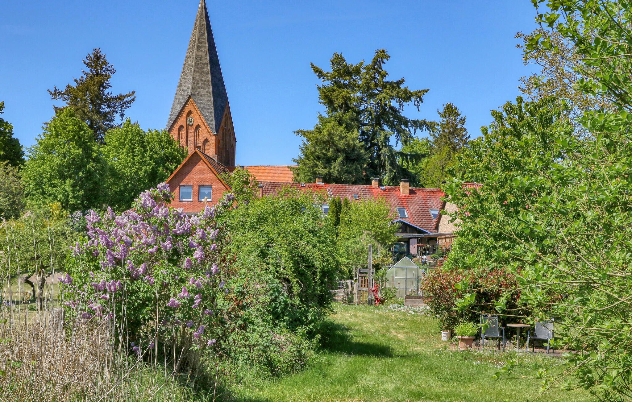 Een kerk met een hoge spits, omringd door weelderige begroeiing en bloeiende planten.