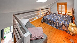 Een slaapkamer in een loft met een houten vloer, een bed met quilt, een raam met jaloezieën en een lamp.