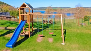A playground with a slide and swings, surrounded by a fenced area and a lake in the background. - Ferienwohnung Neuhausen (Neuhausen/Erzgeb.)