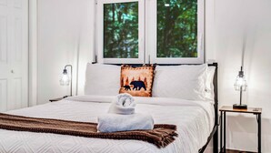 Un lit bien fait, avec une serviette pliée, un plaid marron et un oreiller orné d’un motif d’ours. On y trouve deux lampes de chevet et une fenêtre donnant sur des arbres.