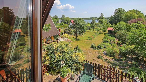 Uitzicht vanaf een balkon op een aangelegde tuin met een meer, met een houten hek, een stoel en een klein gebouw met een rood dak.