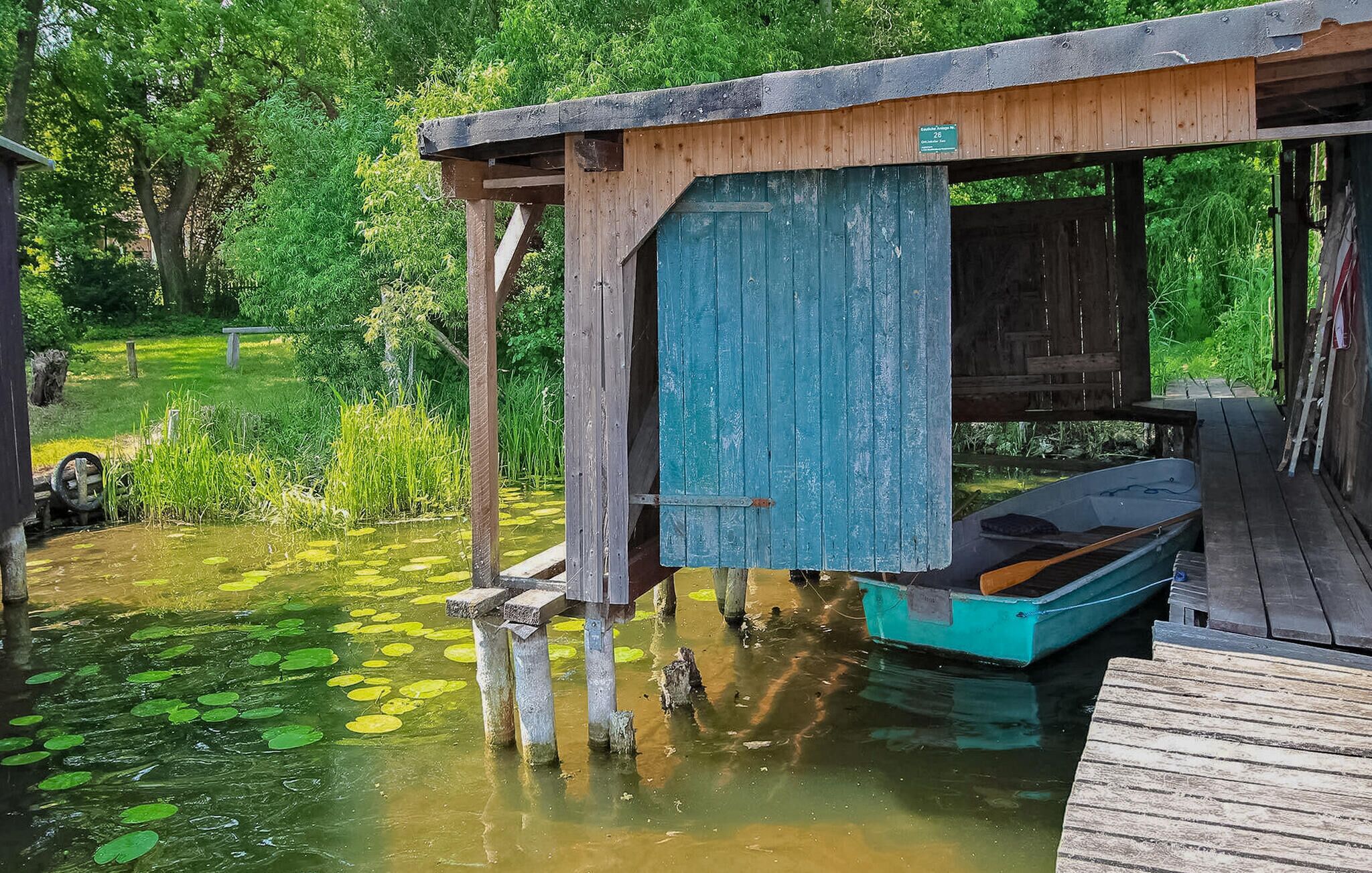 A rustic wooden dock extending into a pond with lily pads, a small boat, and a wooden structure with a blue door.