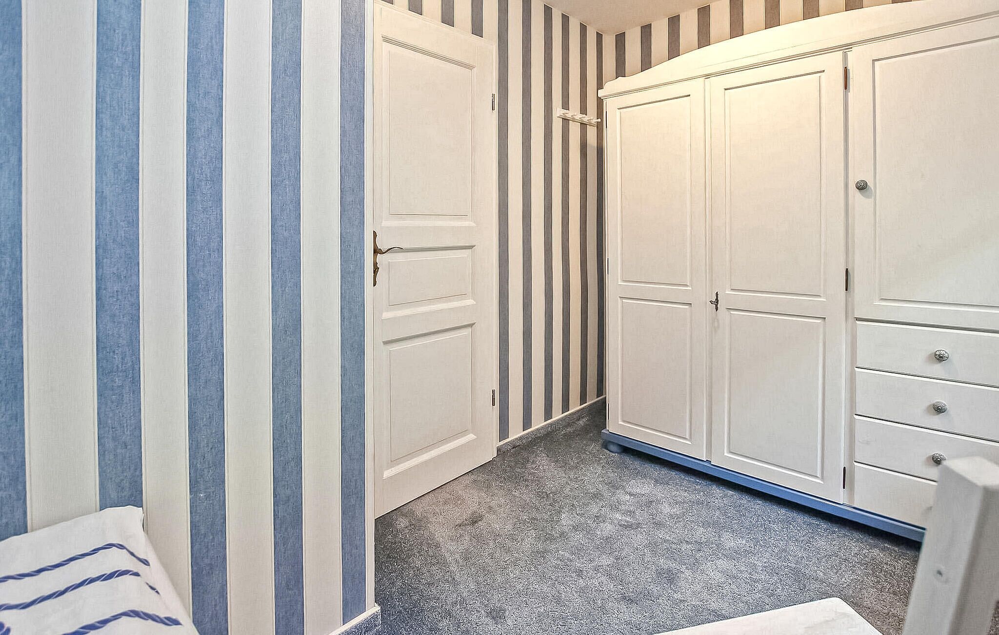 A narrow hallway with striped wallpaper, a white door, and a white wardrobe with drawers.