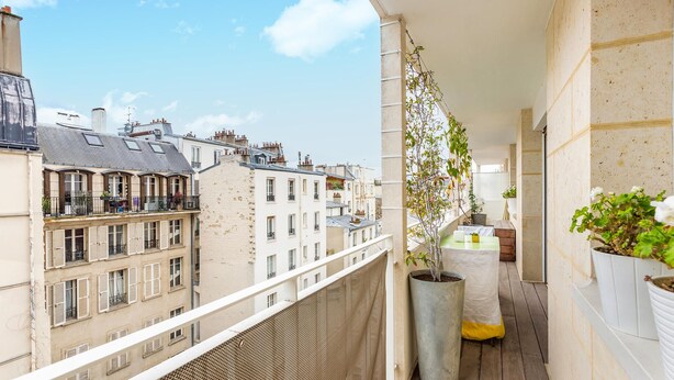 Un balcon étroit donnant sur les bâtiments voisins, une grande plante en pot et une petite table.