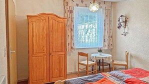 Ein Schlafzimmer mit einem Holzkleiderschrank, einem Bett mit farbenfroher Steppdecke, einem kleinen runden Tisch mit Stuhl und einem Fenster mit gemusterten Vorhängen.