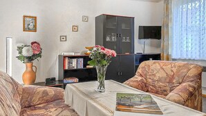 Ein Wohnzimmer mit einer Couch, einem Couchtisch mit einem Buch, einem Bücherregal, einem Fernseher und einem Fenster mit Vorhängen.