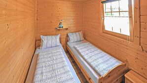A small wooden cabin room with two single beds, a small shelf with a lamp, and a window with a view of greenery.