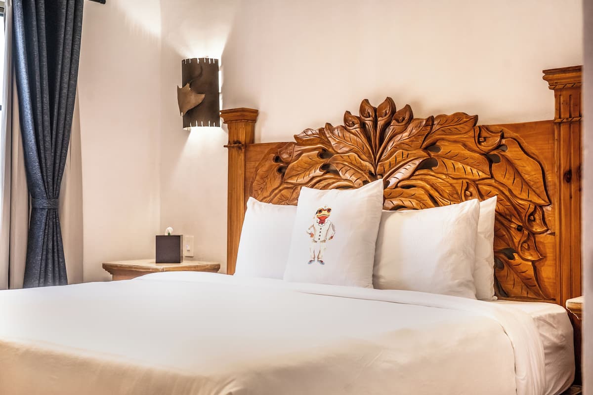 Ein Hotelzimmer mit einem großen Bett, einem hölzernen Kopfende mit Blattfriesen, weißer Bettwäsche und einem kleinen Nachttisch mit Lampe.