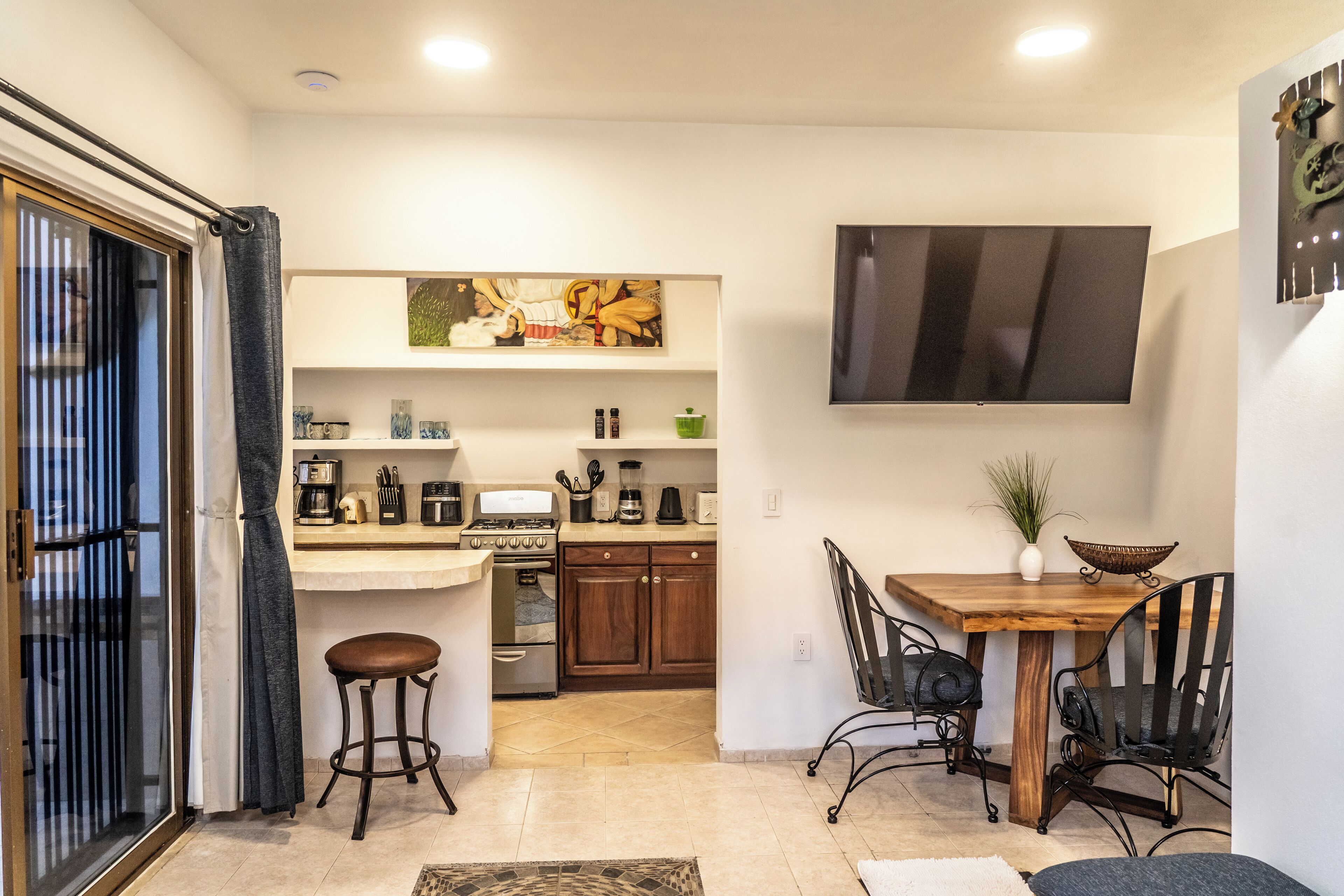 A modern kitchen with wooden cabinets, a dining table with metal chairs, a flat-screen TV, and a sliding glass door leading to a balcony.