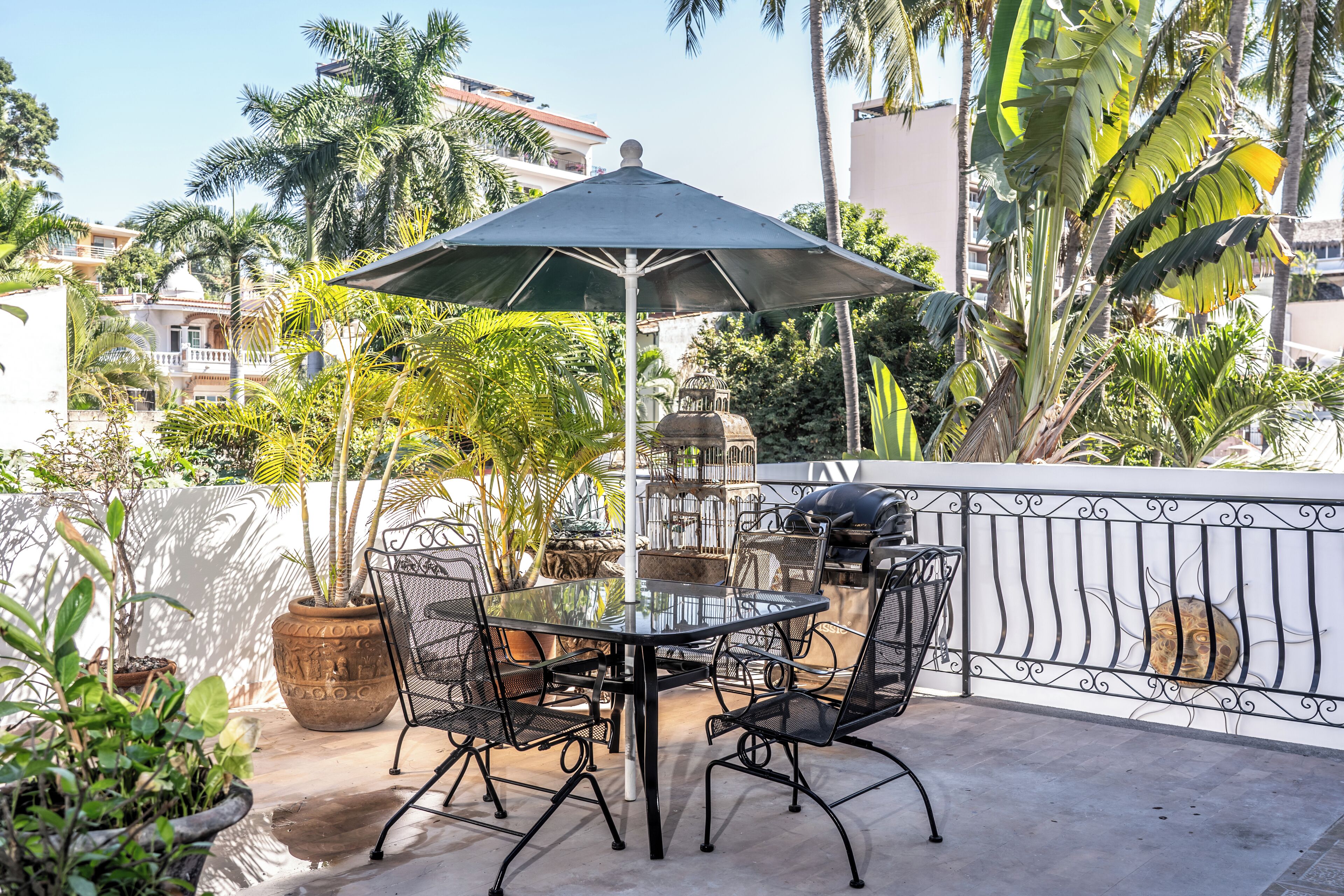 Een buitenpatio met een glazen tafel en stoelen, omringd door plantenpotten en een decoratieve hek.