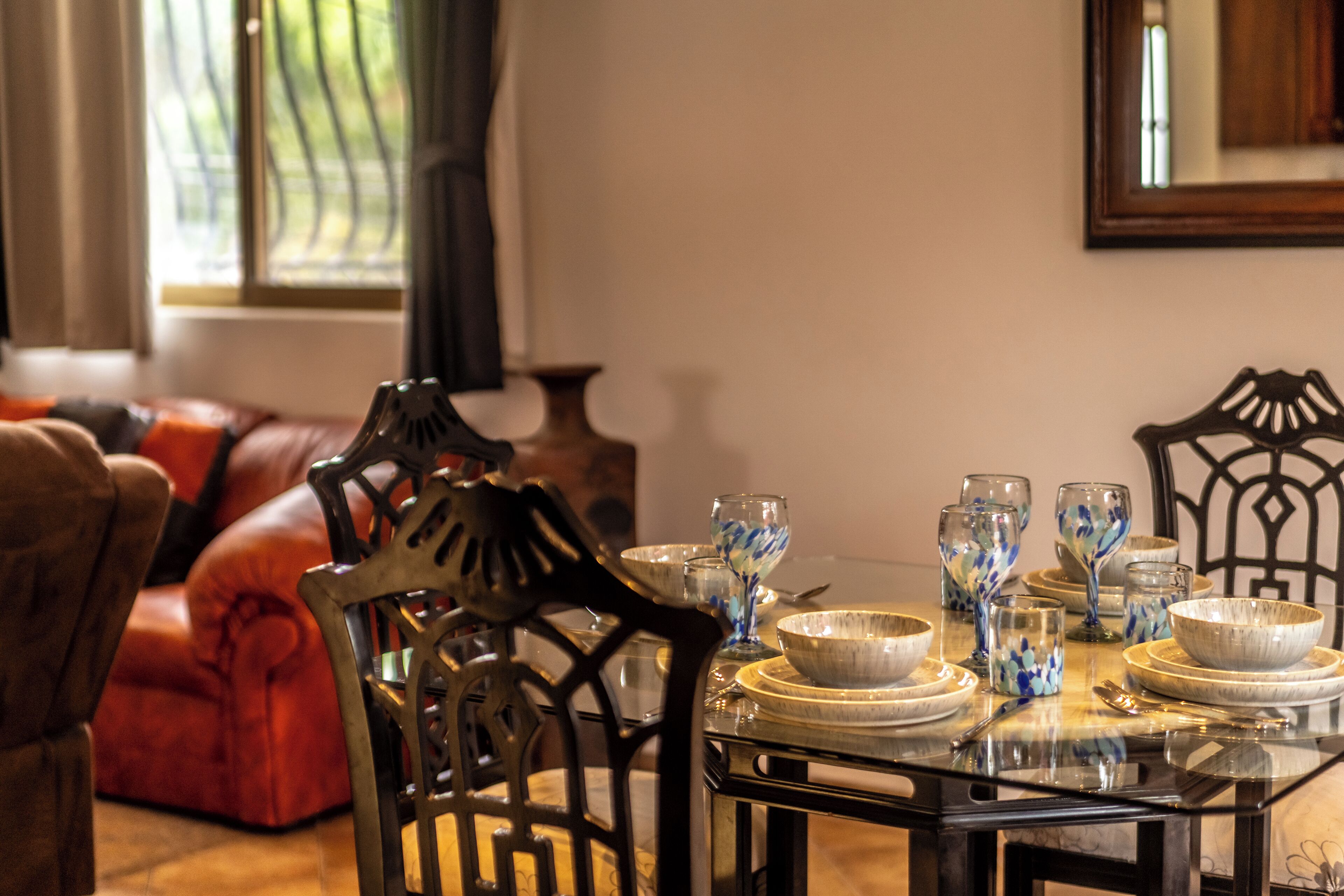 A dining table set with glassware and bowls, surrounded by chairs and a sofa in a well-lit room with a window.