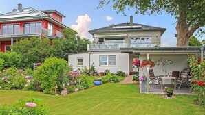 Une maison à deux étages dotée d’un balcon, entourée d’un jardin bien entretenu où poussent diverses plantes et fleurs.