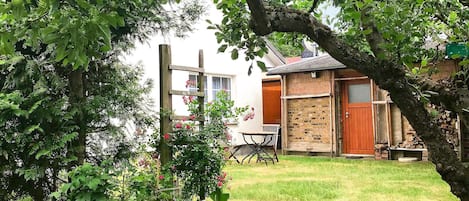 Un jardí amb una taula i caires de fusta, un edifici de rajol amb una porta vermella i una paret blanca amb una finestra.