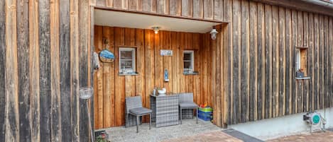 A rustic wooden building with a covered patio area featuring two chairs and a table, a small window, and a door.