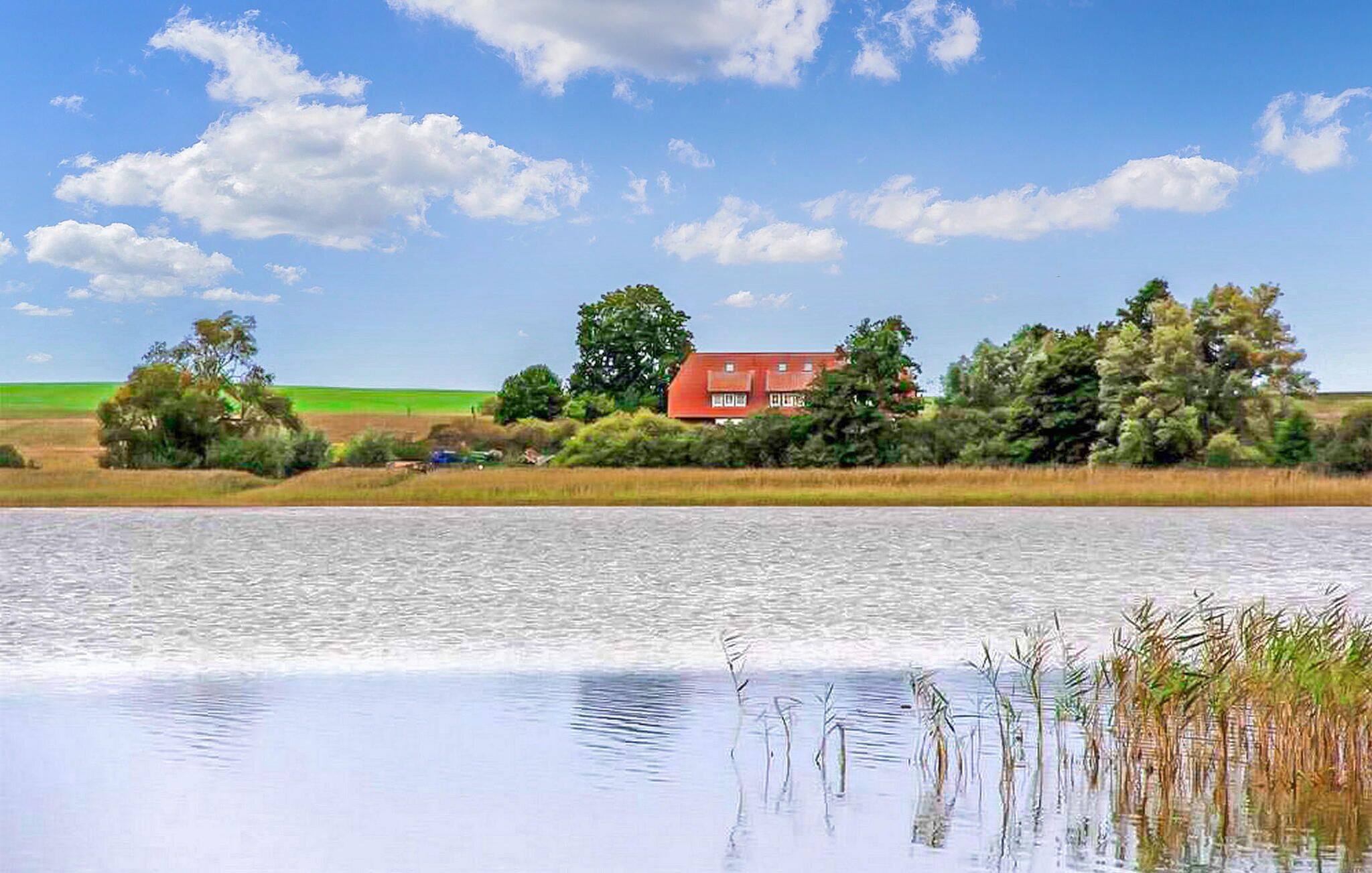 A house by a calm lake with a red roof, surrounded by trees and open fields.