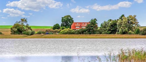 Una casa junto a un lago tranquilo con techo rojo, rodeada de árboles y campos abiertos.