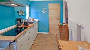 Une cuisine de taille réduite avec des meubles blancs, une cuisinière noire et un plan de travail en bois. On y trouve une chaise en osier et une table avec un verre de vin.