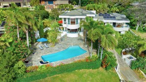 A modern two-story house with a swimming pool, surrounded by lush greenery and palm trees.