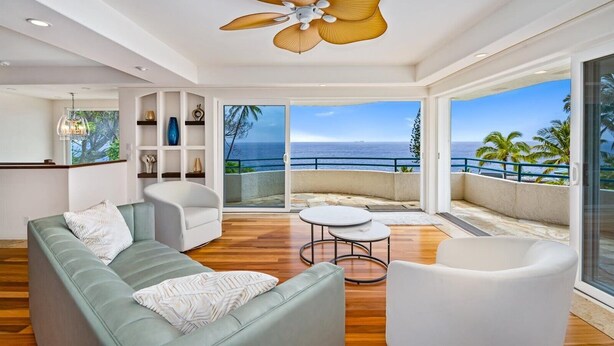 A modern living room with a large sofa, two armchairs, a coffee table, and a ceiling fan. There is a sliding glass door leading to a balcony with a view of the ocean.