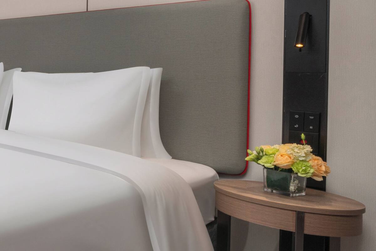 A neatly made bed with white linens, a small round table with a vase of flowers, and a wall-mounted light fixture.