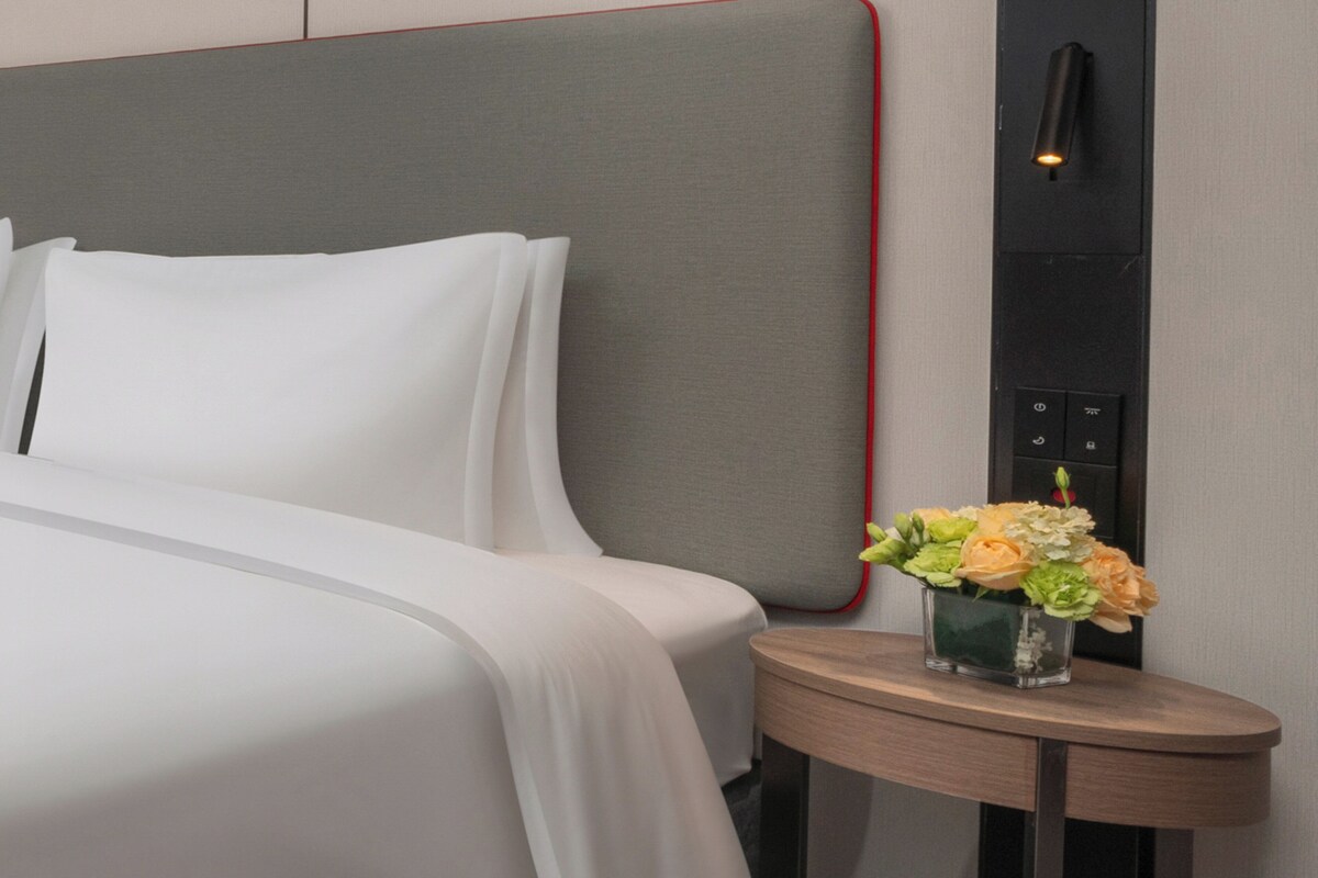 A neatly made bed with white linens, a small round table with a vase of flowers, and a wall-mounted light fixture.