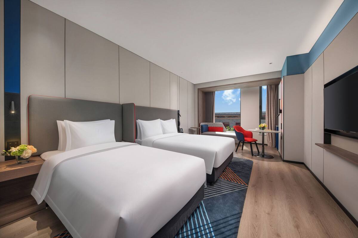 A modern hotel room with two beds, a TV, a desk, and a view of the city.