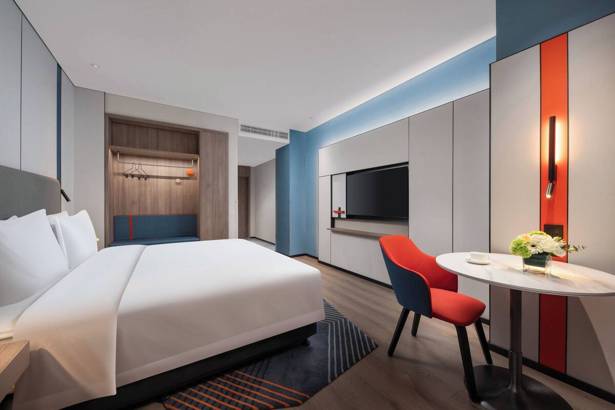 A modern hotel room with a large bed, a TV, a small table with a vase of flowers, and a seating area with a red chair.