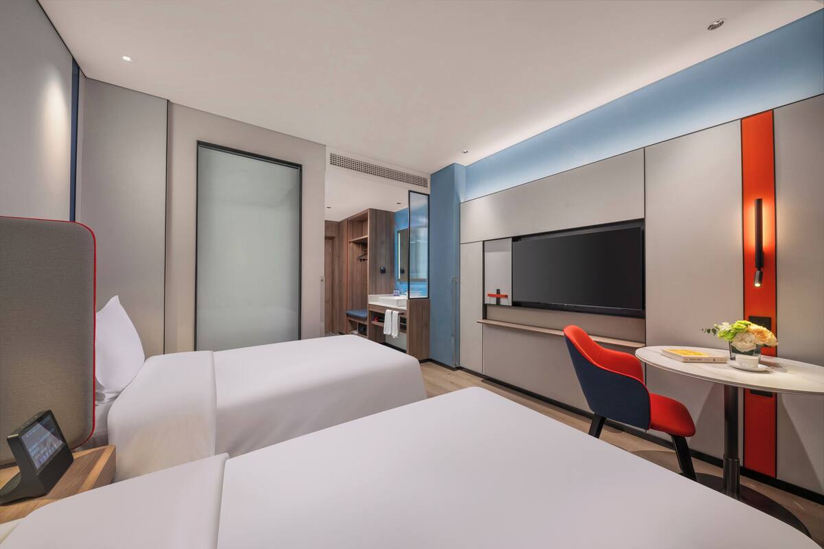 A hotel room with two beds, a TV, a small table with a vase of flowers, and a desk with a computer.