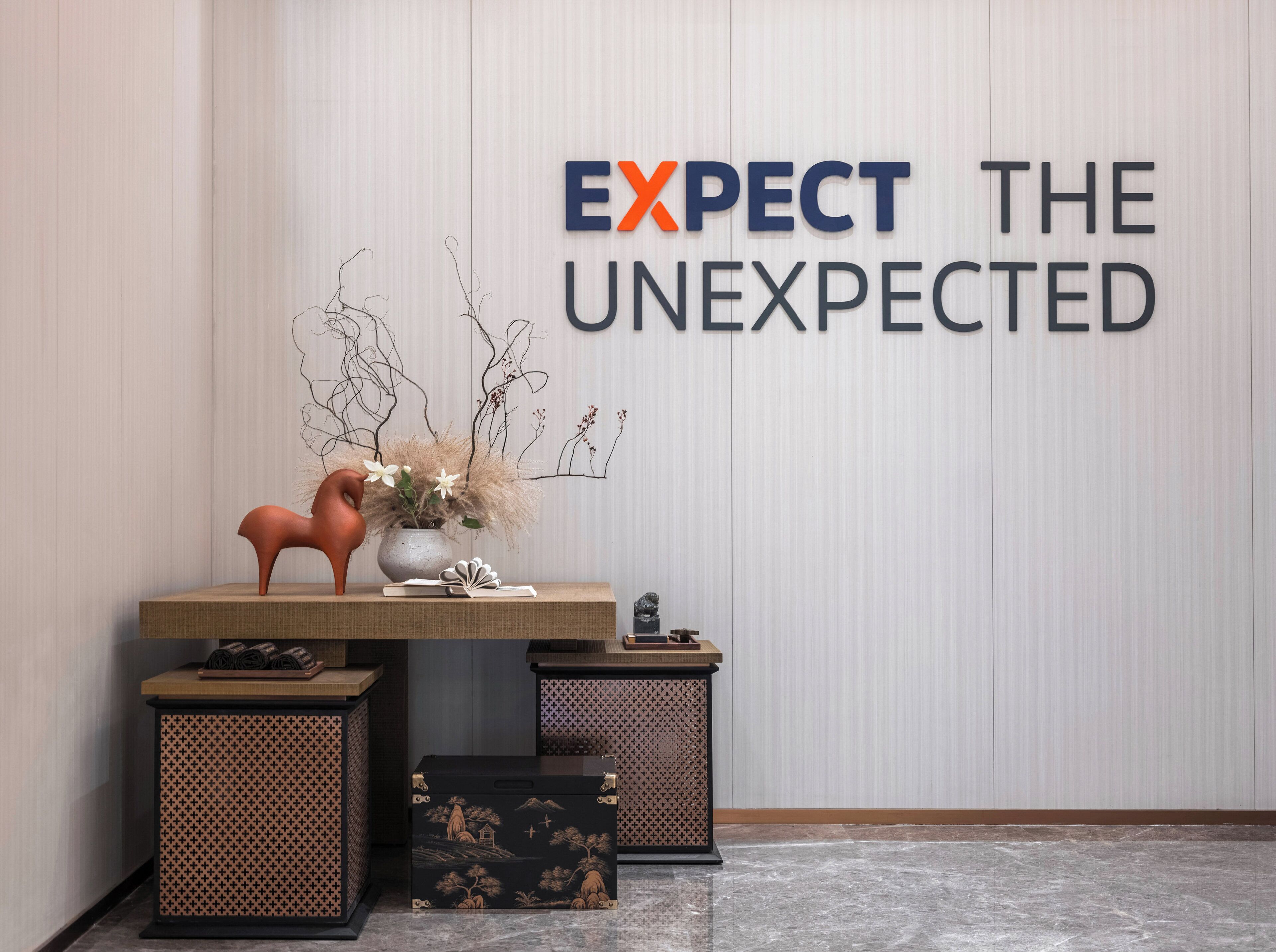 A reception area with a wall sign reading "EXPECT THE UNEXPECTED," a wooden table with a vase of flowers, a decorative horse sculpture, and two side tables with patterned boxes.