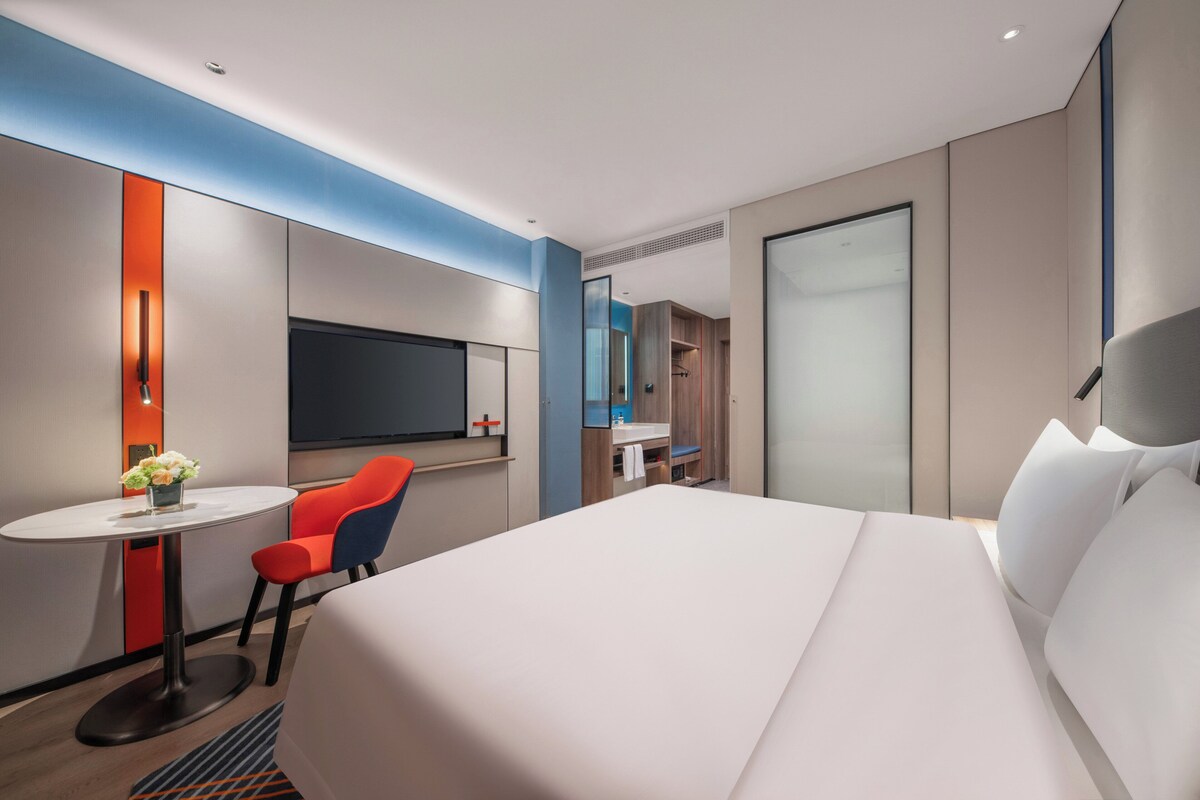 A modern hotel room with a large bed, a desk with a chair, a small table with a vase of flowers, a TV, and a bathroom with a mirror and sink.
