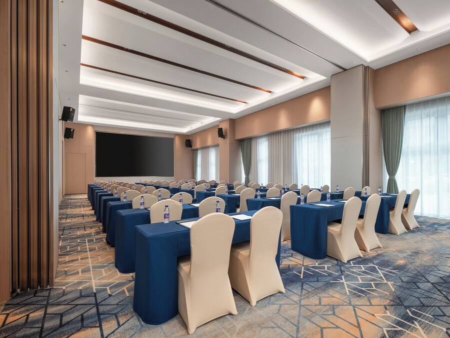 A conference room with rows of tables and chairs, a large screen, and a carpeted floor.